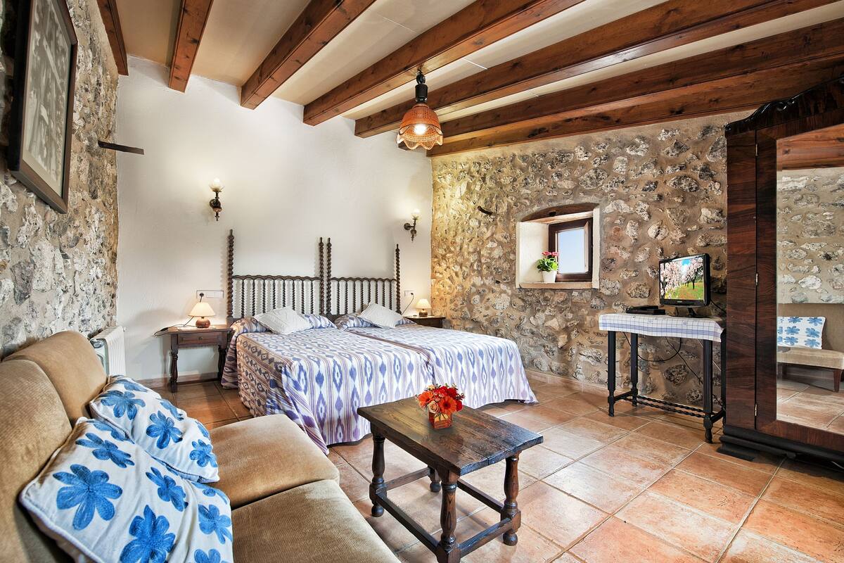 A rustic room with a bed, wooden beams, stone walls, a small table with a vase, and a television.