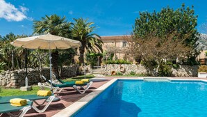 A swimming pool with lounge chairs and umbrellas, surrounded by greenery and a stone wall. - Can Guillo (Pollenca)