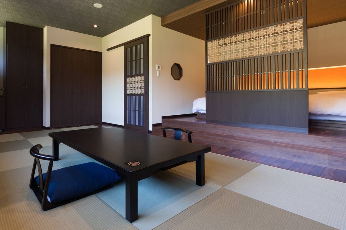 A traditional Japanese-style room with tatami flooring, a low table with cushions, a bed with a wooden headboard, and a sliding door with a decorative screen.