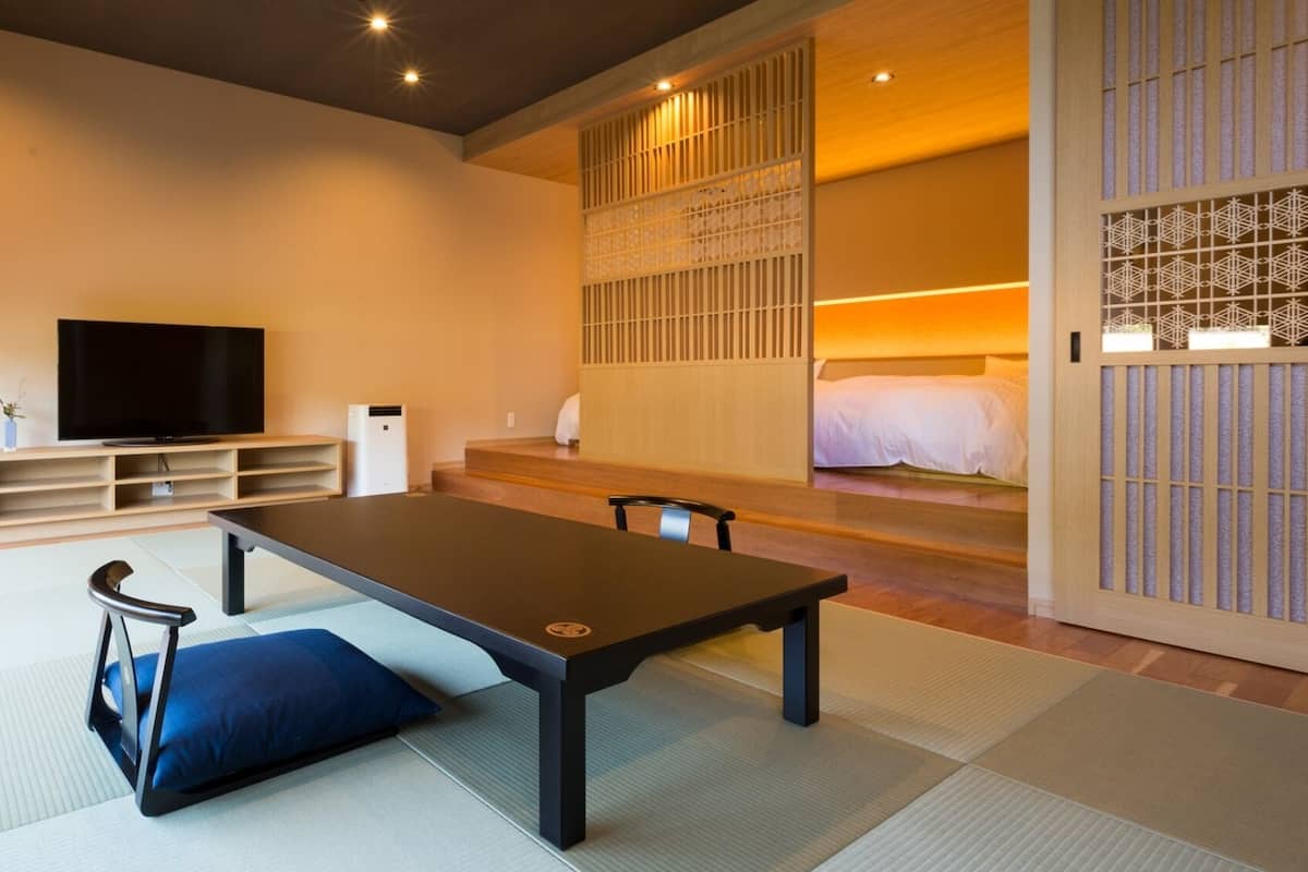A modern room with a tatami mat floor, a low table, a TV, and a bed with a wooden screen.