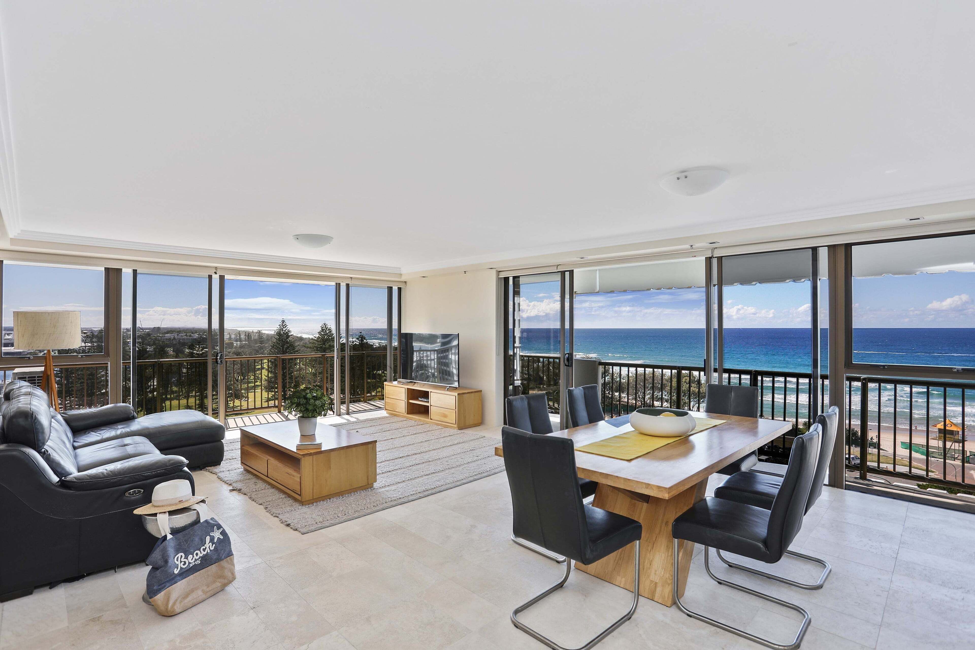 A spacious living room with a balcony overlooking the ocean, furnished with a sofa, coffee table, and dining area.