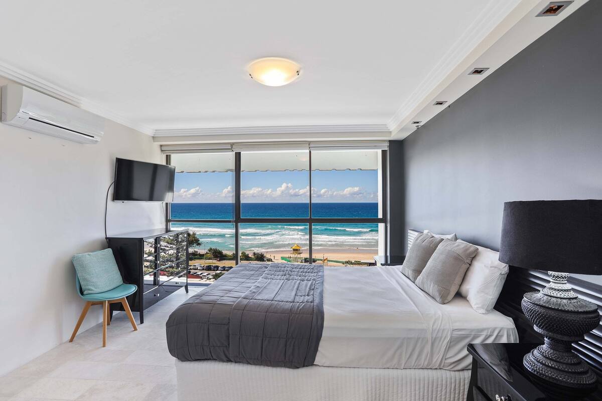 Ein Schlafzimmer mit großem Fenster mit Blick auf einen Strand, einem Bett mit weißen Leinentüchern, einem schwarzen Nachttisch, einem Türkis-Sessel und einer schwarzen Lampe.