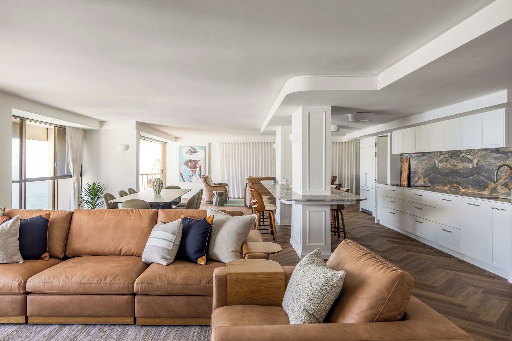 A modern living room with a brown leather sofa, a dining area with a white table, and a kitchen with white cabinets and a marble backsplash.