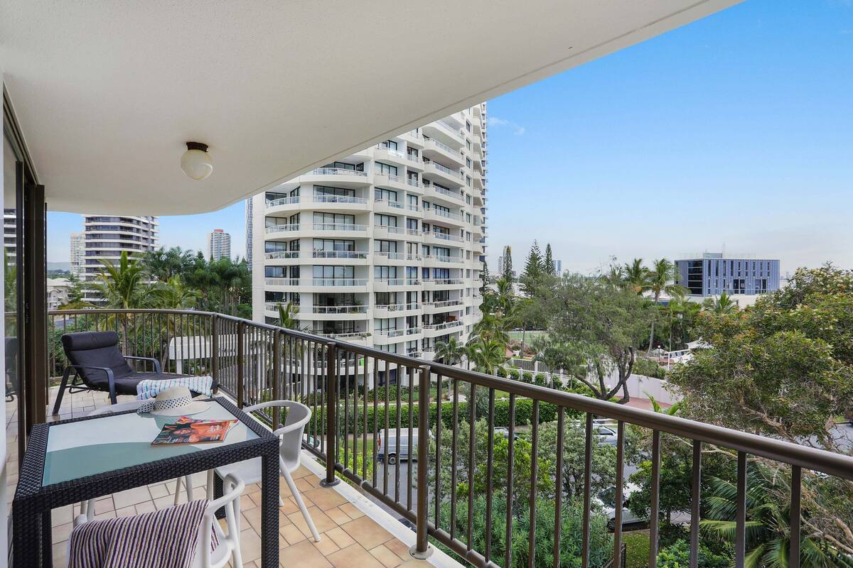 Ein Balkon mit Glastisch, Stühlen und Blick auf ein modernes Gebäude sowie die umliegende Grünanlage.