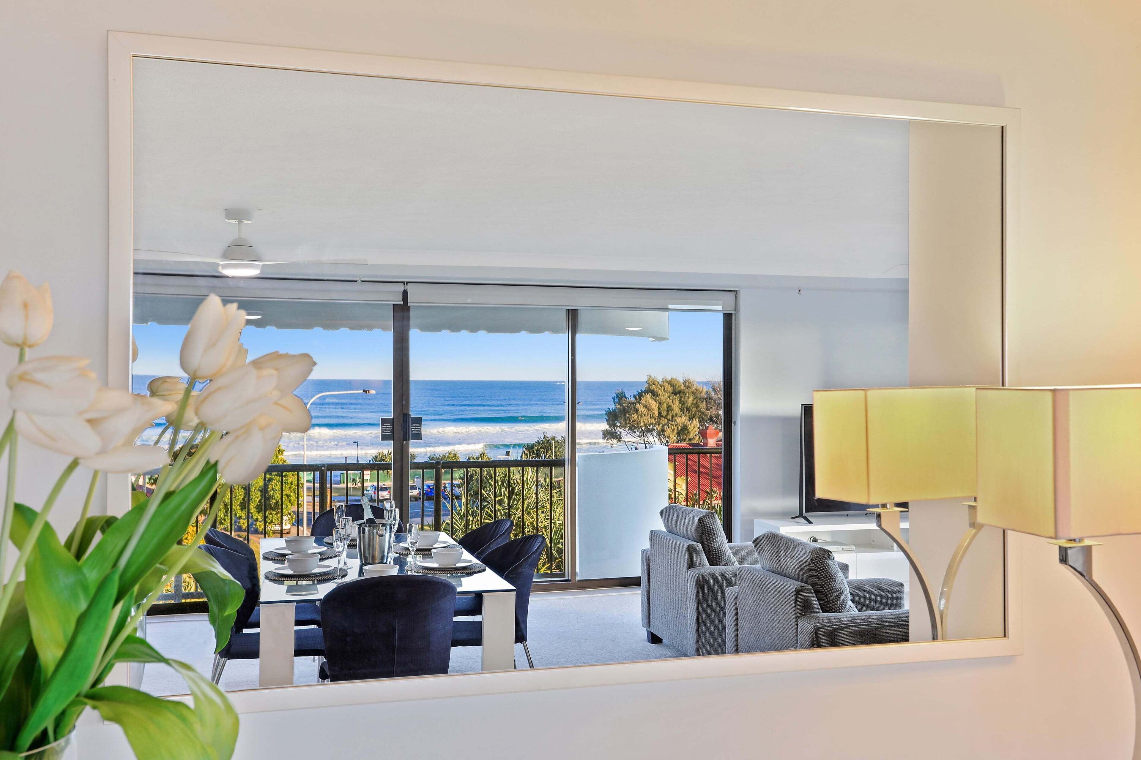 A modern room with a balcony overlooking the sea, a dining area with a table set, and a sofa.