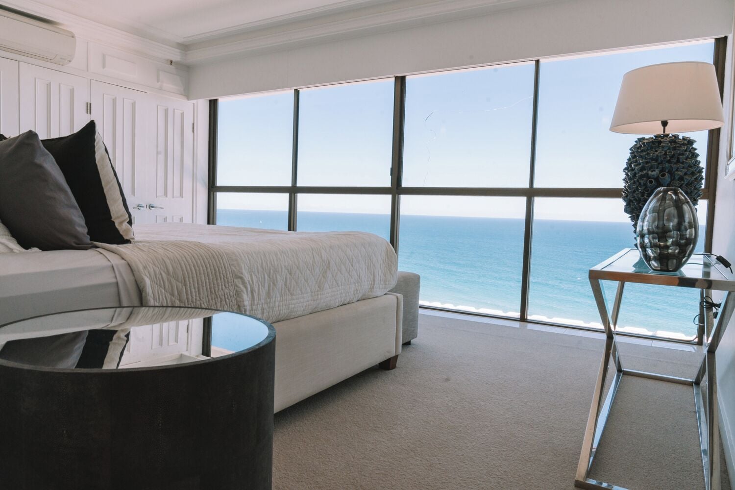 A bedroom with a large window overlooking the ocean, a bed with a white headboard, a round table with a lamp, and a decorative vase.