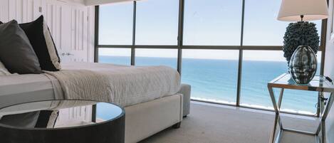 A bedroom with a large window overlooking the ocean, a bed with a white headboard, a round table with a lamp, and a decorative vase.