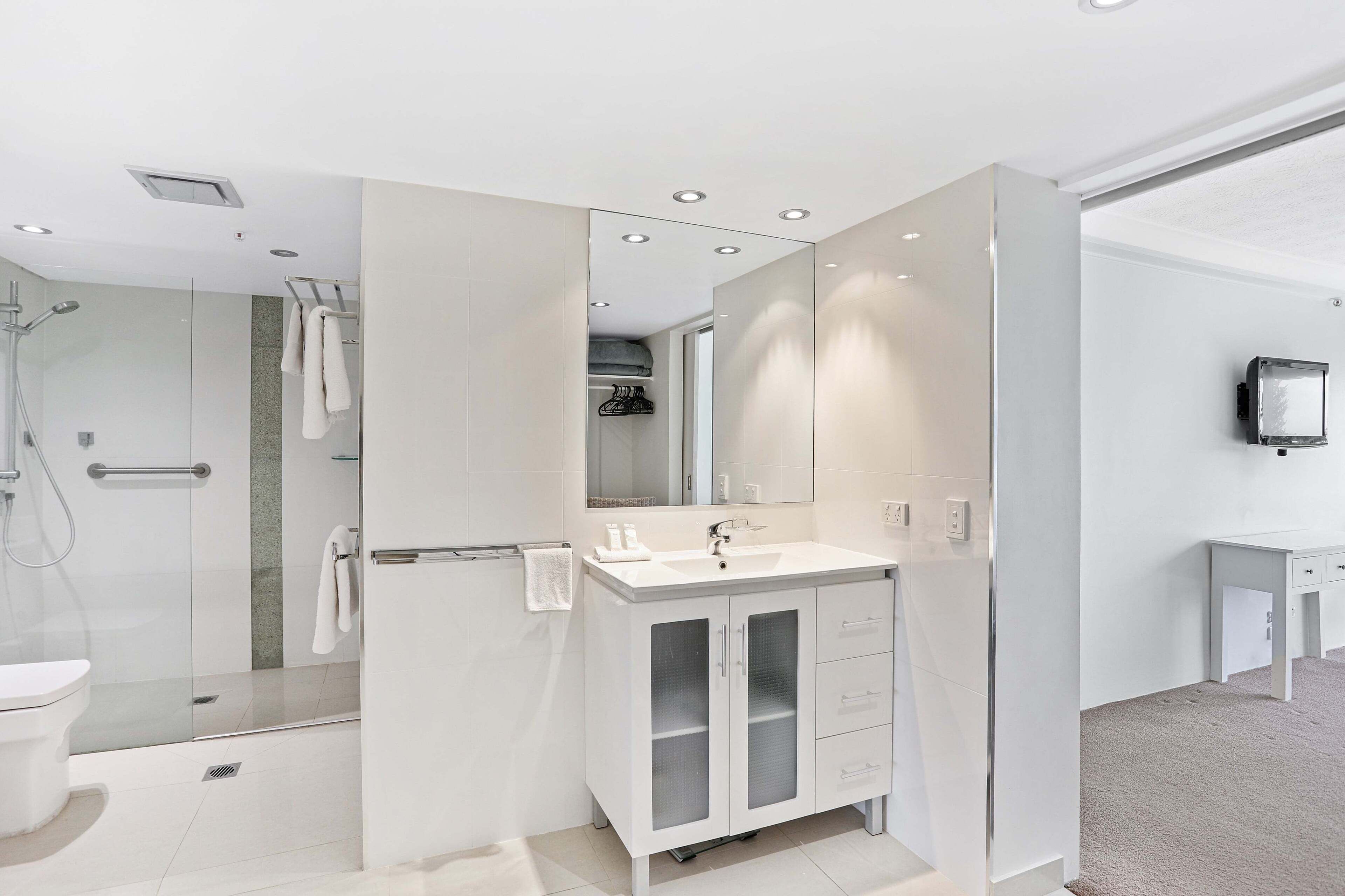 Ein modernes Badezimmer mit Duschkabine aus Glas, einem weißen Waschbecken mit Spiegel darüber und einem kleinen Schminktisch mit Schubladen.
