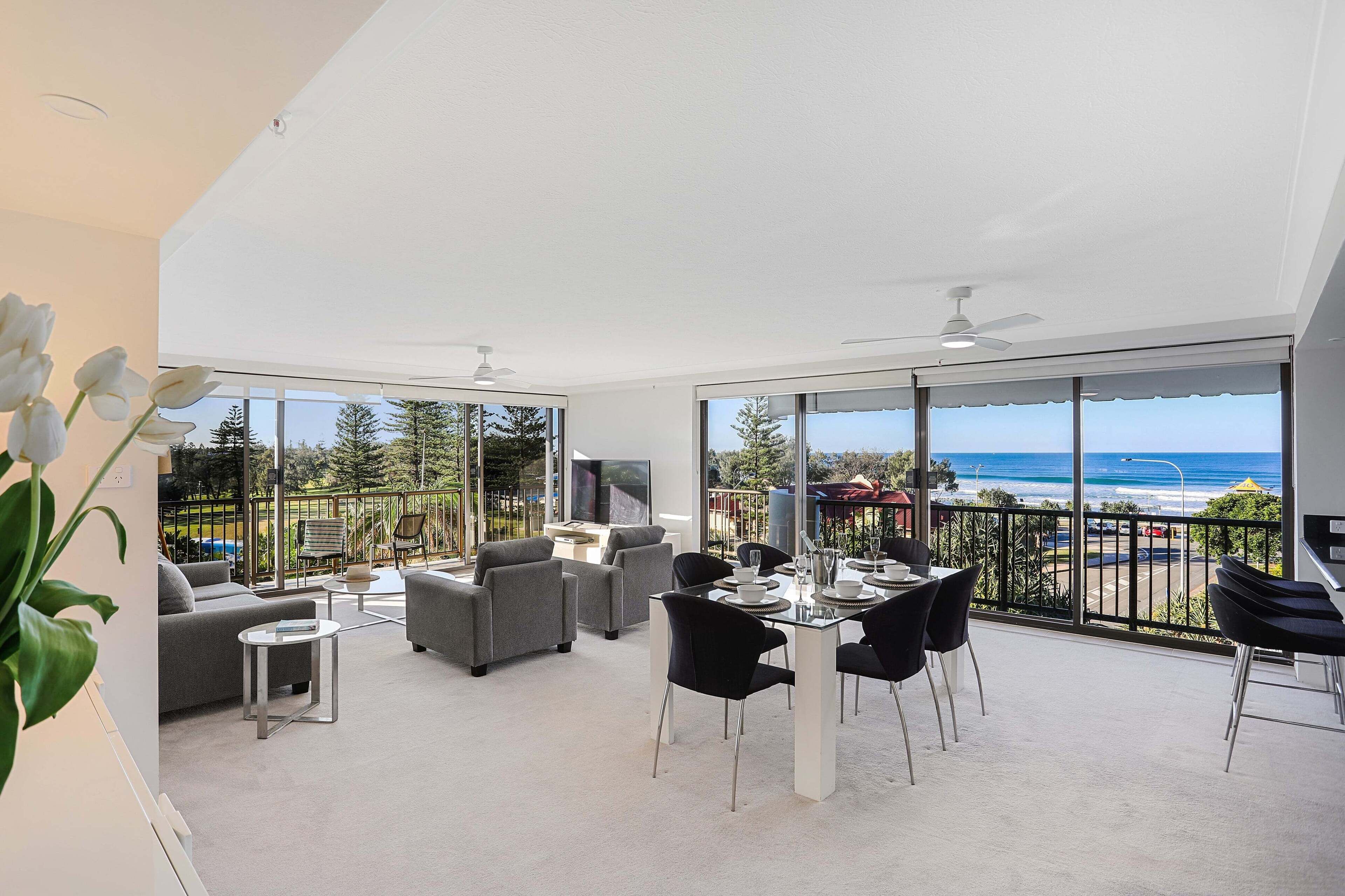 Amplia sala de estar con balcón vista al mar. Hay sofás, una mesa de comedor y un televisor.