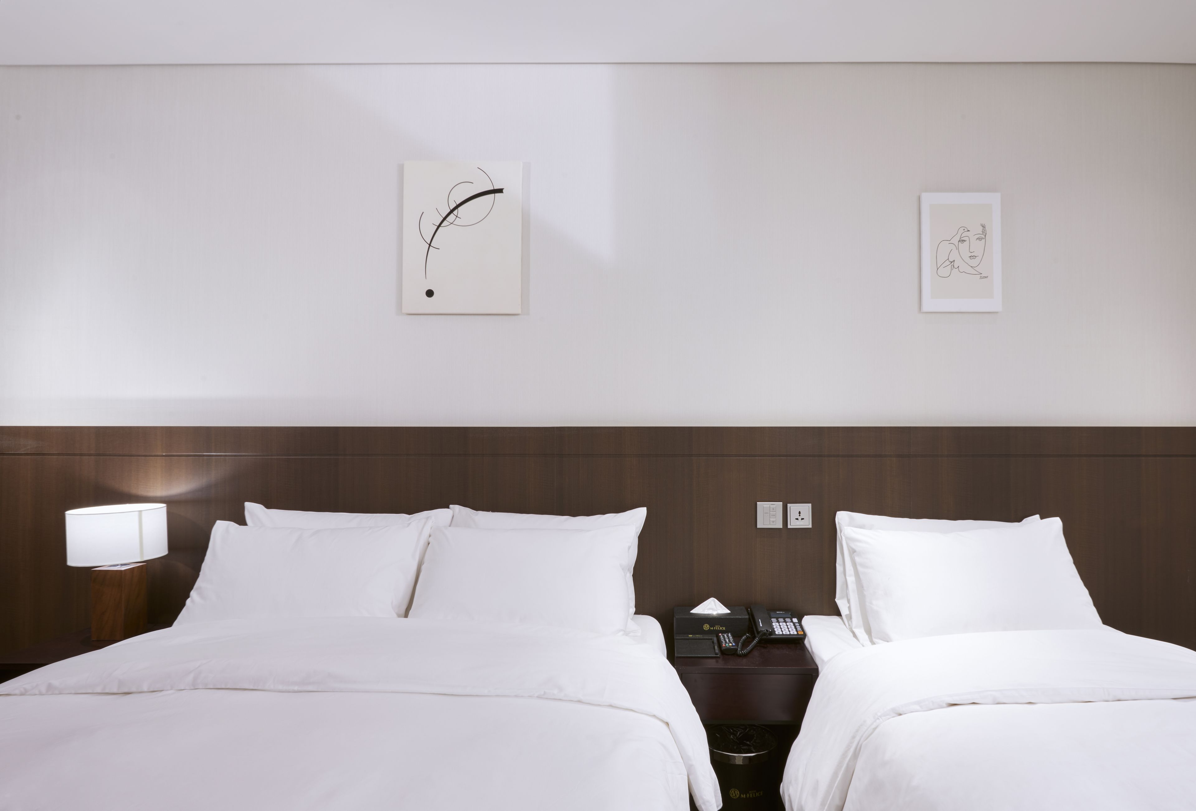A hotel room with two beds, a nightstand with a lamp, a phone, and artwork on the wall.