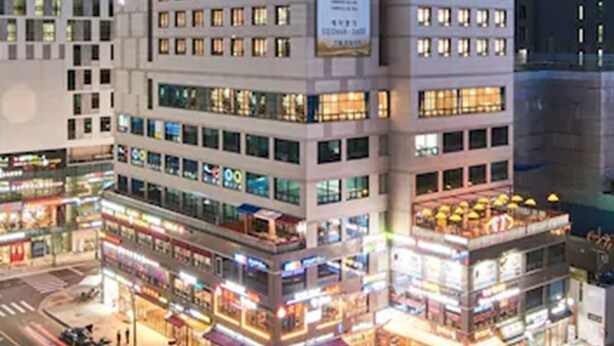 Un hotel de varios pisos con un letrero llamativo, varios locales comerciales en la planta baja y una concurrida calle urbana.