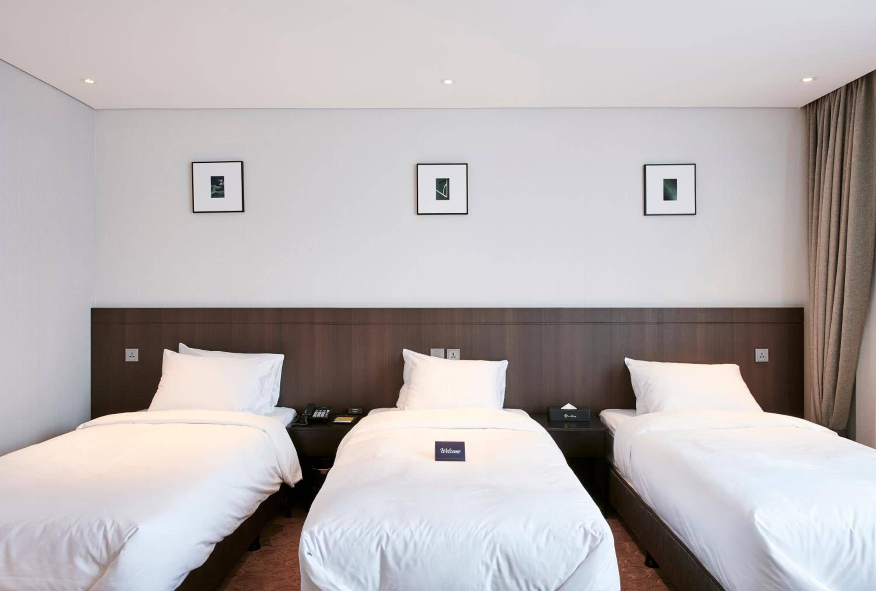 A hotel room with two single beds, a wooden headboard, a nightstand with a phone, and a "Welcome" sign.