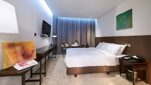Una habitación de hotel con una cama grande, un escritorio con silla, un televisor montado en la pared y un cuadro sobre el escritorio.