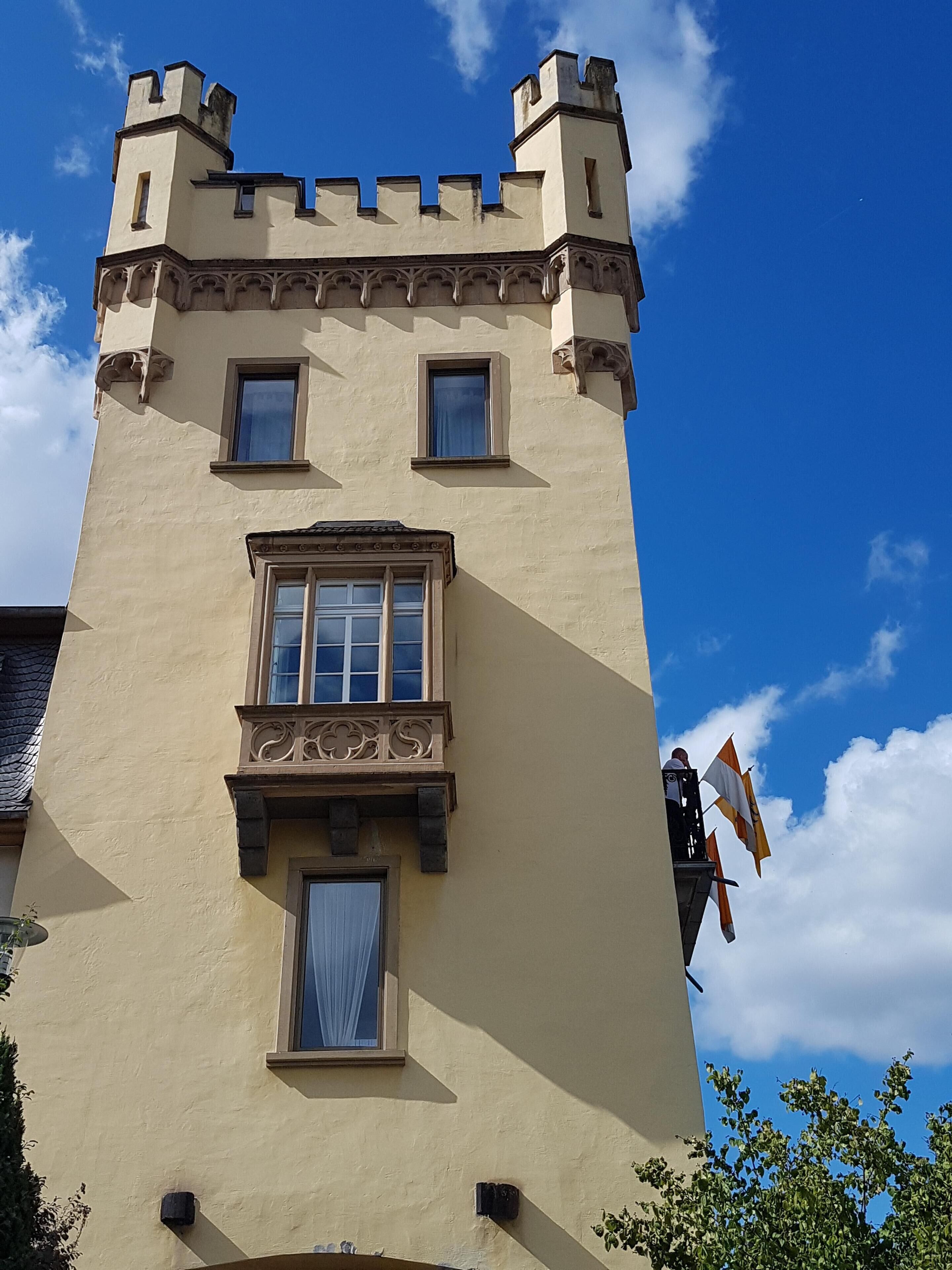 Ein hoch aufragendes, schlossähnliches Gebäude mit einem Türmchen und Zinnen, einem Balkon mit Flaggen und einem Fenster mit Vorhängen.