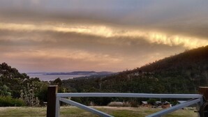 A view of a coastal landscape with a wooden fence in the foreground, a body of water in the middle ground, and a hilly forested area in the background. - Fantastic getaway for family or friends with spectacular views. (Snug)