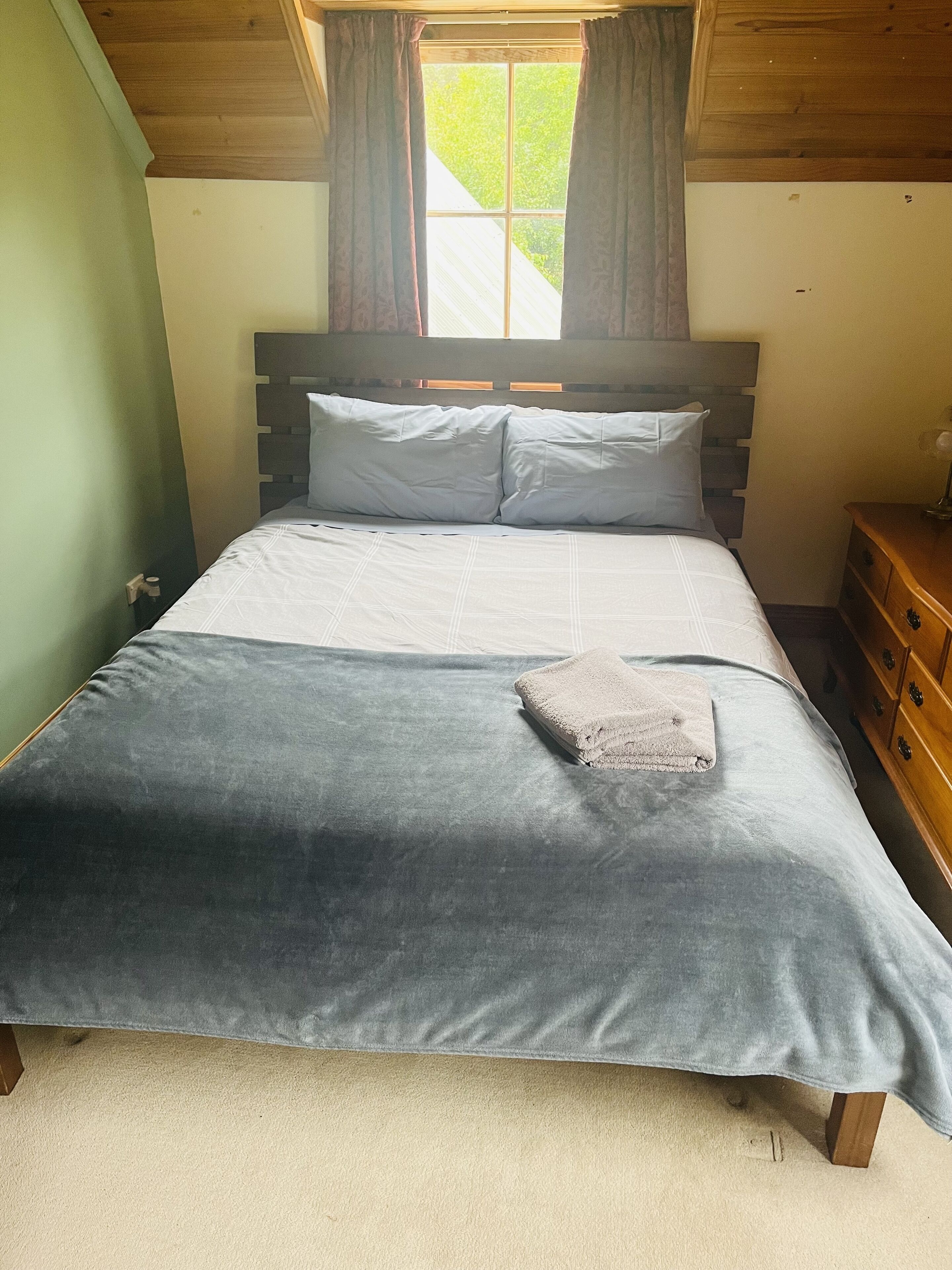 A neatly made bed with a grey duvet and pillows, a wooden nightstand with a lamp, and a window with curtains.