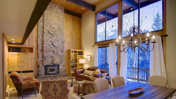 A cozy living room with a stone fireplace, wooden beams, and large windows offering a view of snow-covered trees. - Hike In/Out w/Stunning Mountain Views + Heated Pool-Hot Tub-Gym-Wood Fireplace-W/D in Unit (Steamboat Springs)