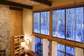 Een gezellige kamer met een stenen muur, houten schappen, een leren bank en grote ramen met uitzicht op een besneeuwd bos.