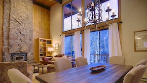 A cozy living room with a stone fireplace, wooden beams, a chandelier, and a large window with a view of trees. - Hike In/Out w/Stunning Mountain Views + Heated Pool-Hot Tub-Gym-Wood Fireplace-W/D in Unit (Steamboat Springs)