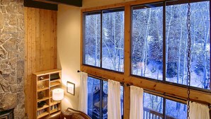 A cozy cabin interior with a stone wall, wooden beams, a fireplace, a bookshelf, and large windows offering a view of a snowy forest. - Hike In/Out w/Stunning Mountain Views + Heated Pool-Hot Tub-Gym-Wood Fireplace-W/D in Unit (Steamboat Springs)