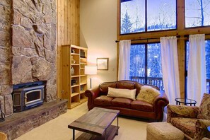 Een gezellige woonkamer met een stenen openhaard, houten planken, een leren bank en een groot raam met uitzicht op met sneeuw bedekte bomen.