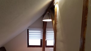 Ein kleines Schlafzimmer unter der Dachschräge mit Holzbalken, einem weißen Teppich, einem braunen Sofa und einem Bett mit einer grün-weiß gestreiften Bettdecke.