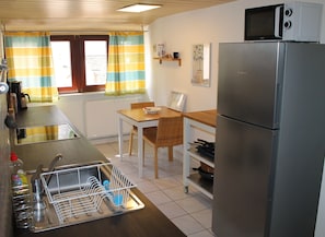 A compact kitchen with a stainless steel refrigerator, a sink with a dish rack, a wooden table with chairs, and a microwave on top of the fridge.