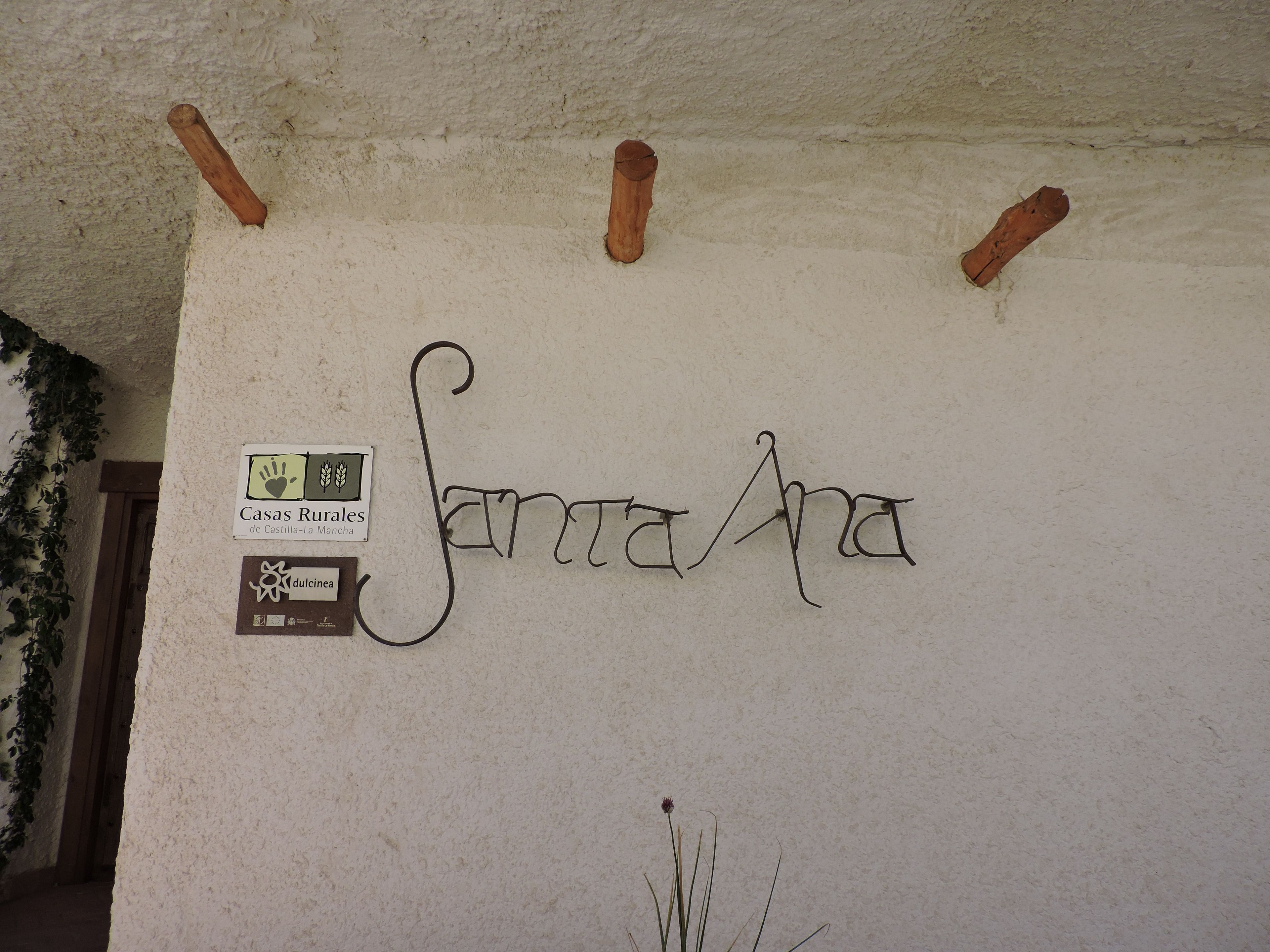 Een muur met de naam 'Santa Ana' in sierlijk schrift, een bordje voor 'Casas Rurales de Castilla-La Mancha', en een bordje met 'dulce de leche'.