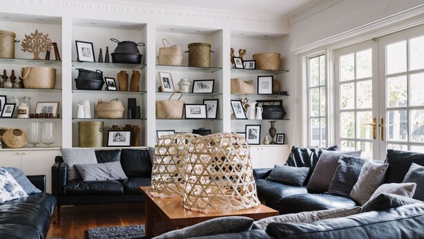 A modern living room with a black leather sofa set, a wooden coffee table, and a large woven basket centerpiece. Shelves above the sofa display various woven baskets and framed pictures.