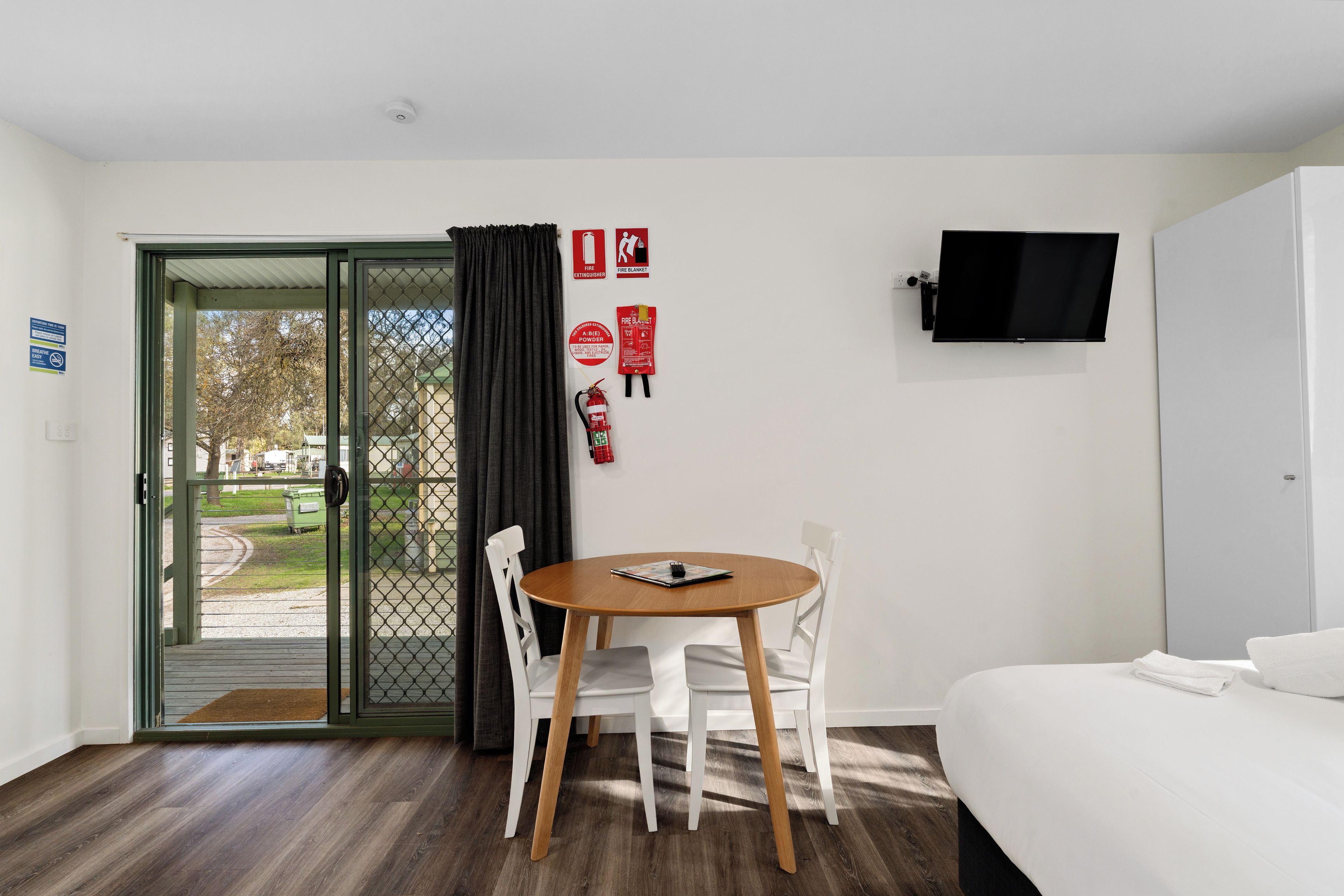 A hotel room with a bed, a TV mounted on the wall, a dining table with chairs, and a door leading outside.