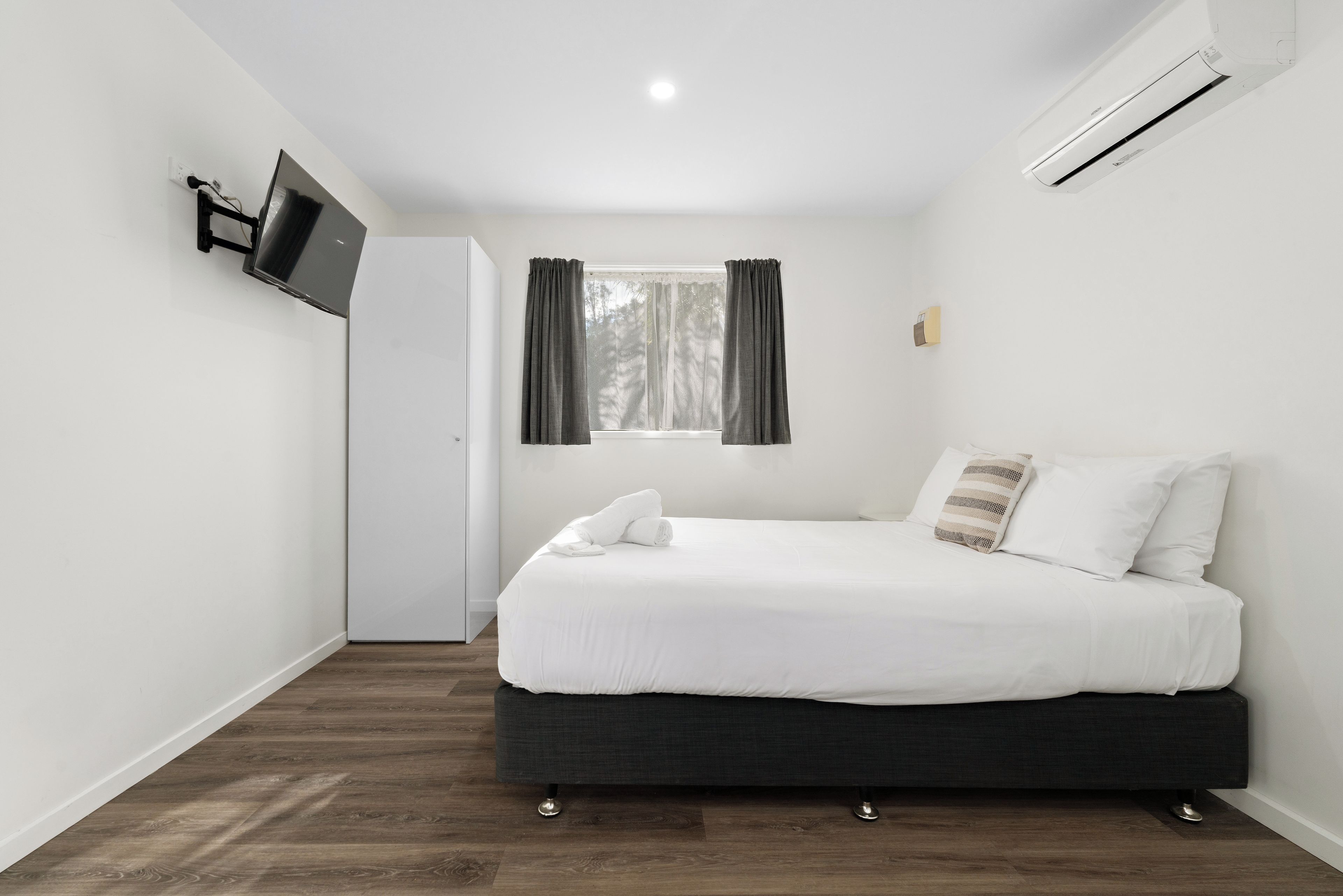A bedroom with a bed, a TV mounted on the wall, a window with curtains, and an air conditioning unit.