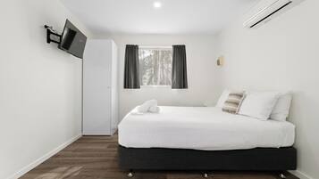 A bedroom with a bed, a TV mounted on the wall, a window with curtains, and an air conditioning unit.