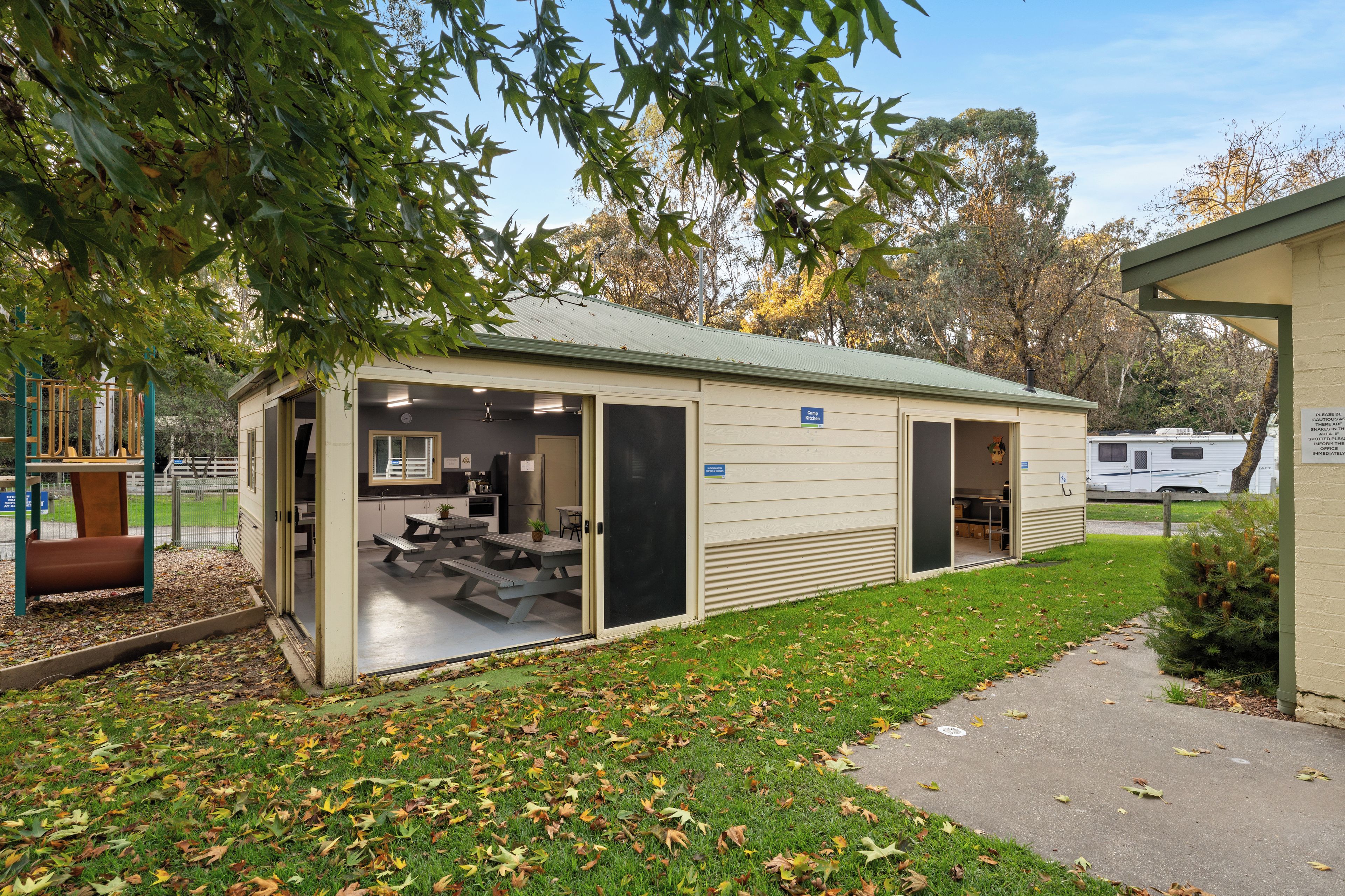 A small, single-story building with a kitchen area visible through an open door, picnic tables outside, and a sign indicating a camp kitchen.