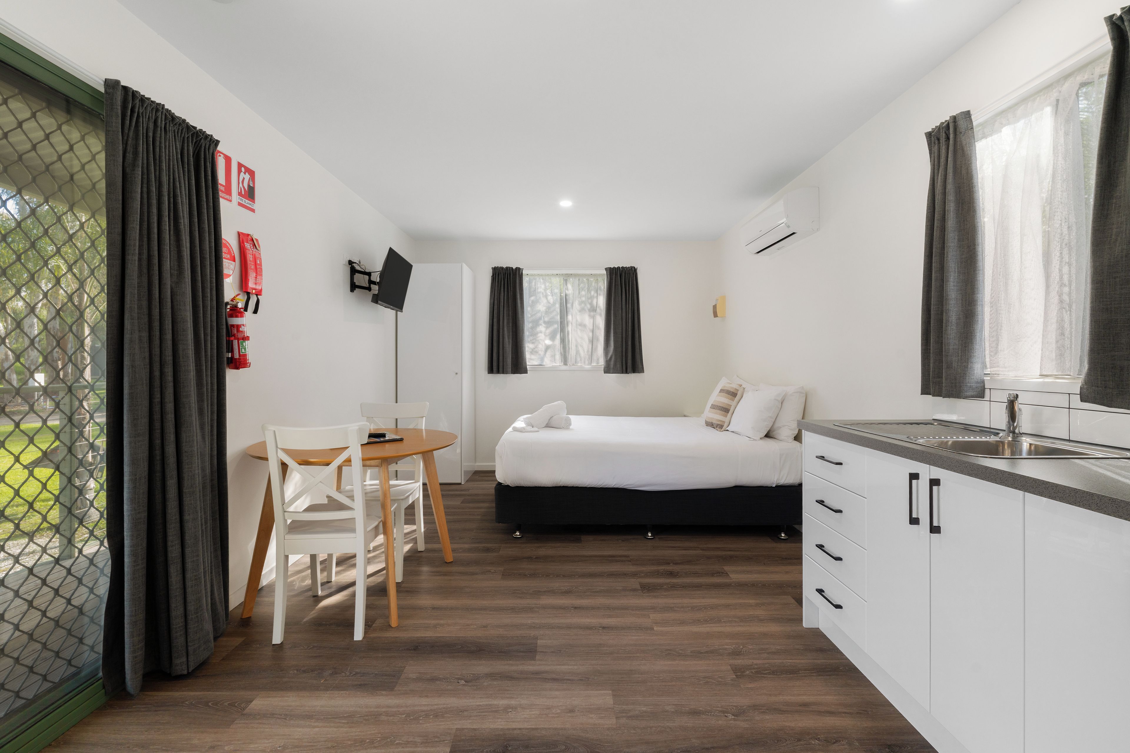 Habitación de hotel compacta con cama, una pequeña mesa de comedor con sillas, kitchenette y ventana con cortinas.