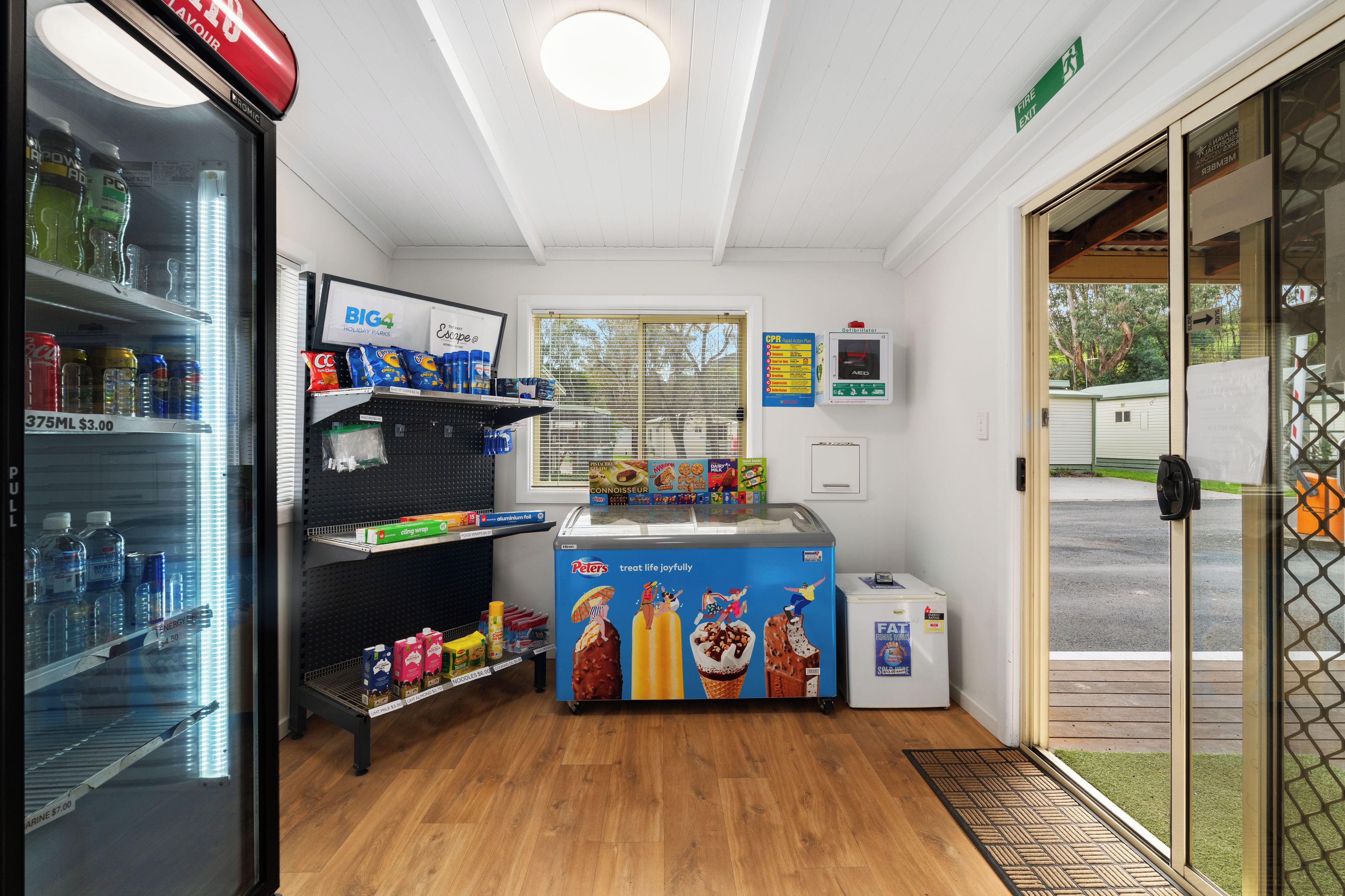 A small ice cream shop with a freezer, shelves stocked with snacks, and a window with blinds.