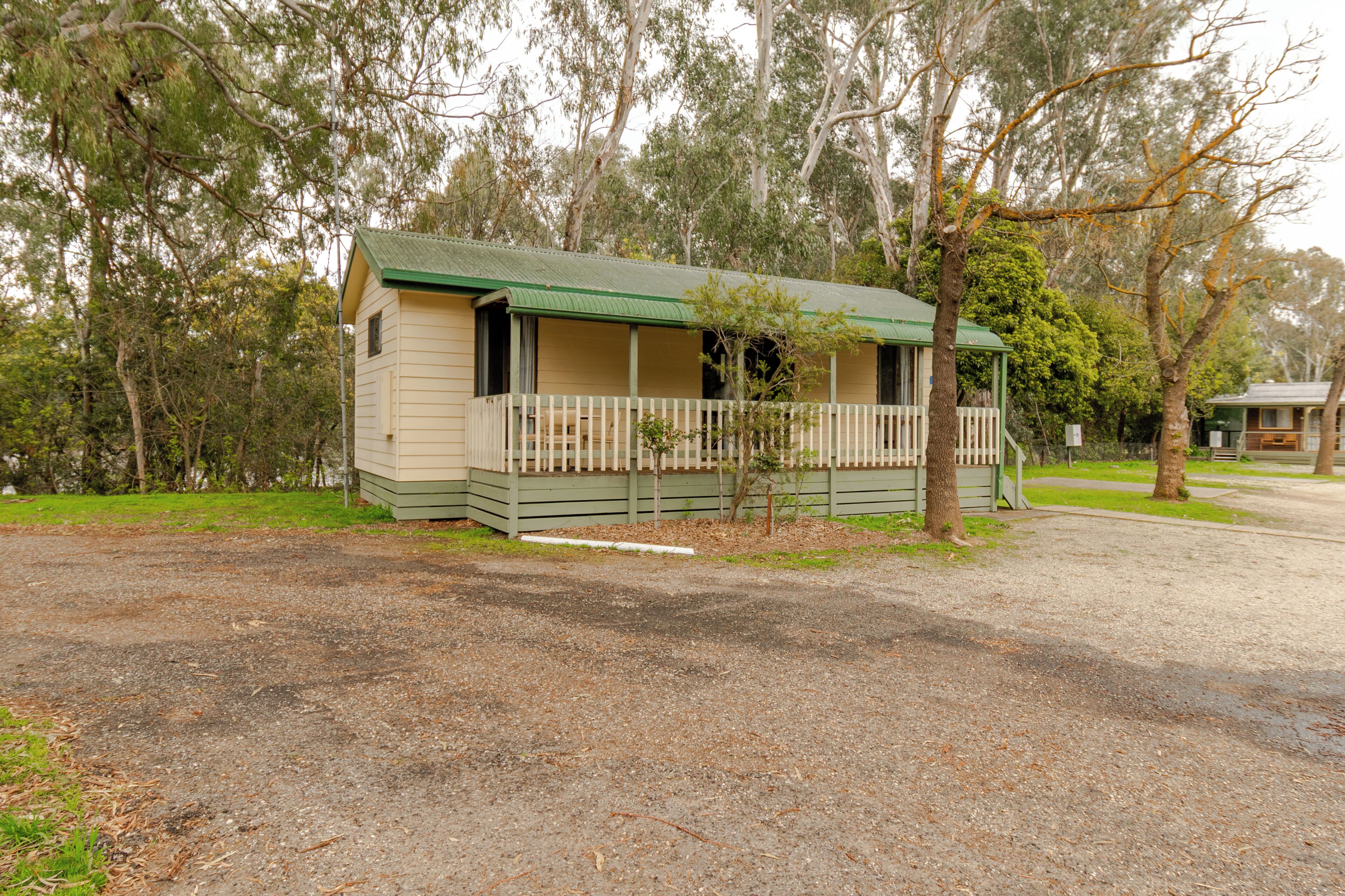 A small, single-story building with a green roof, a porch with a white railing, and a gravel driveway.