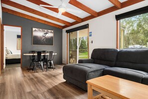 Un salon moderne avec un canapé gris foncé, une table basse en bois et un coin repas avec des chaises en métal. On peut apercevoir un ventilateur de plafond, et une grande fenêtre offre une vue sur les arbres.