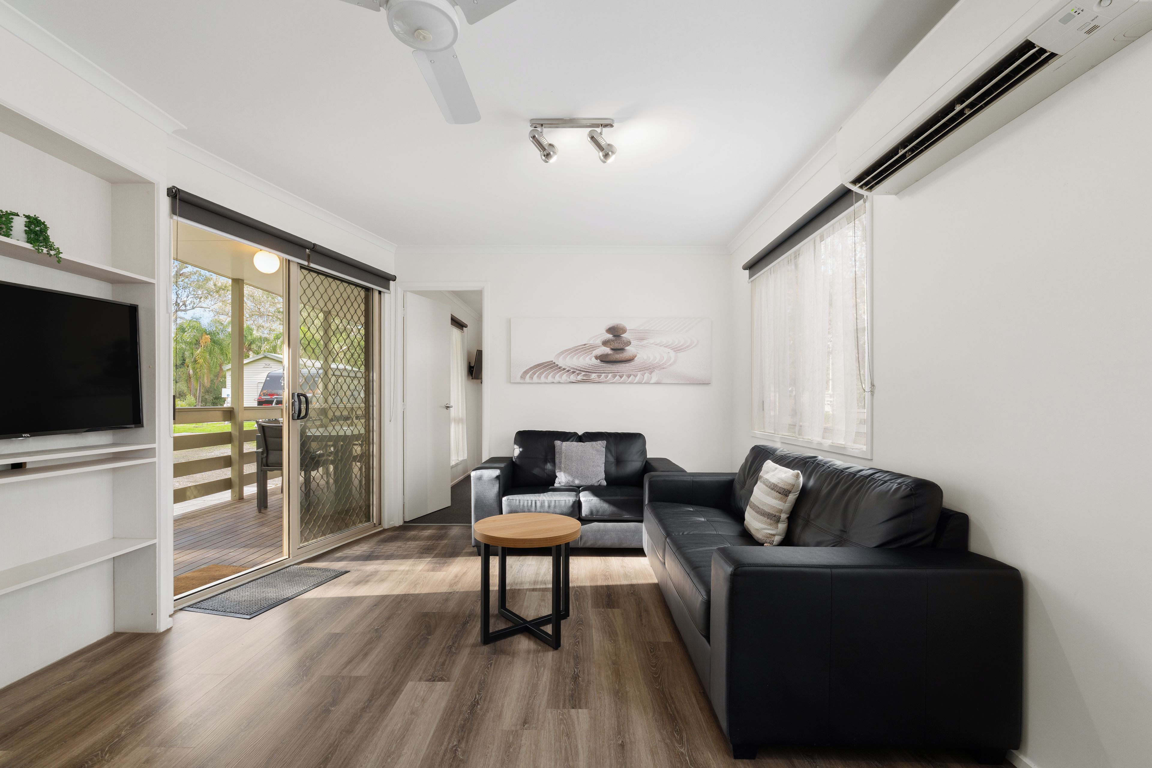 A modern living room with a flat-screen TV, a black leather sofa, a wooden coffee table, and a sliding glass door leading to an outdoor area.