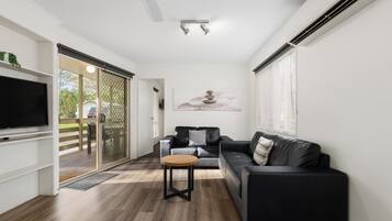 A modern living room with a flat-screen TV, a black leather sofa, a wooden coffee table, and a sliding glass door leading to an outdoor area.