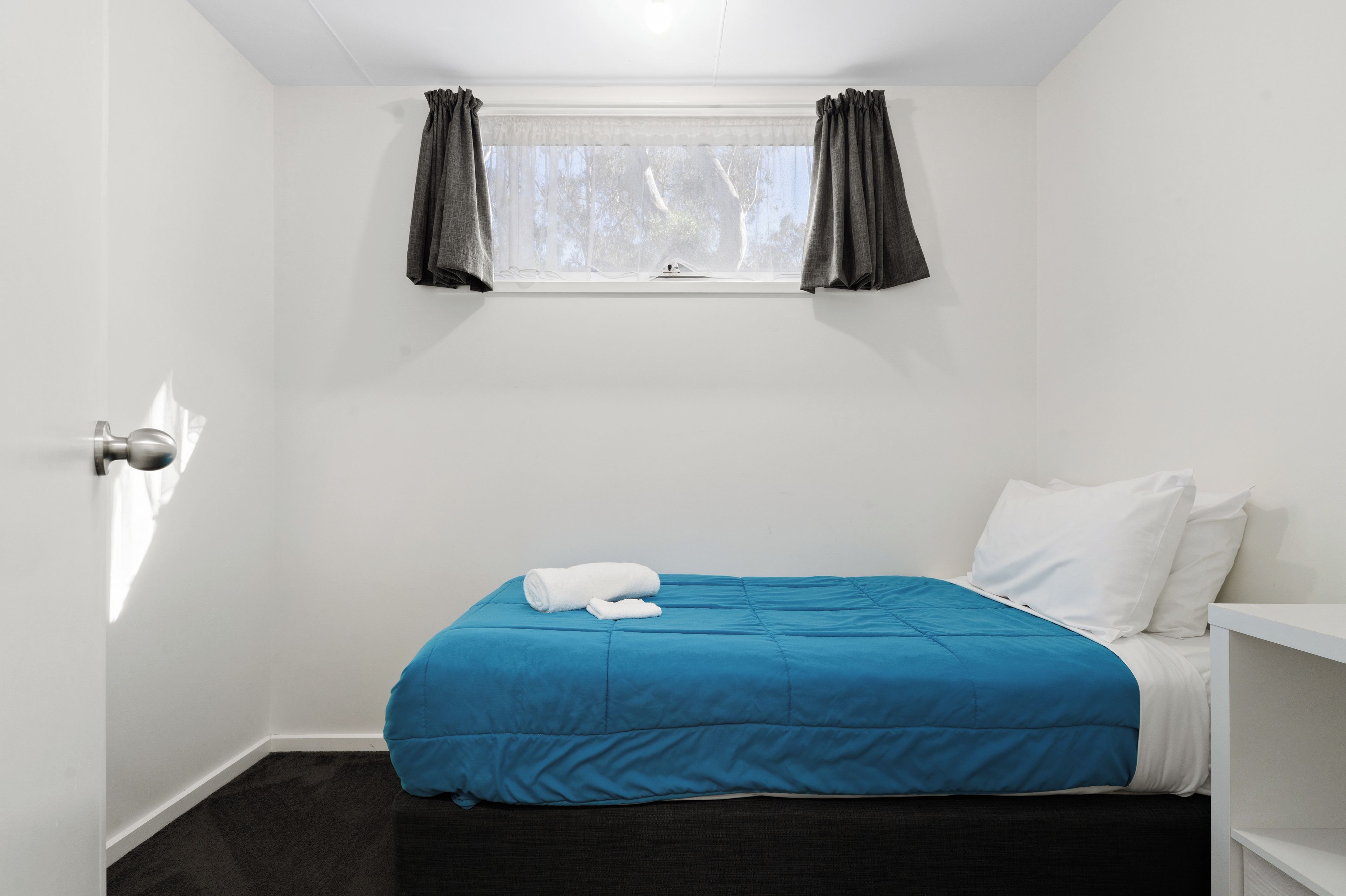 A small, single-bed room with a window, a white door, and a dark carpet.