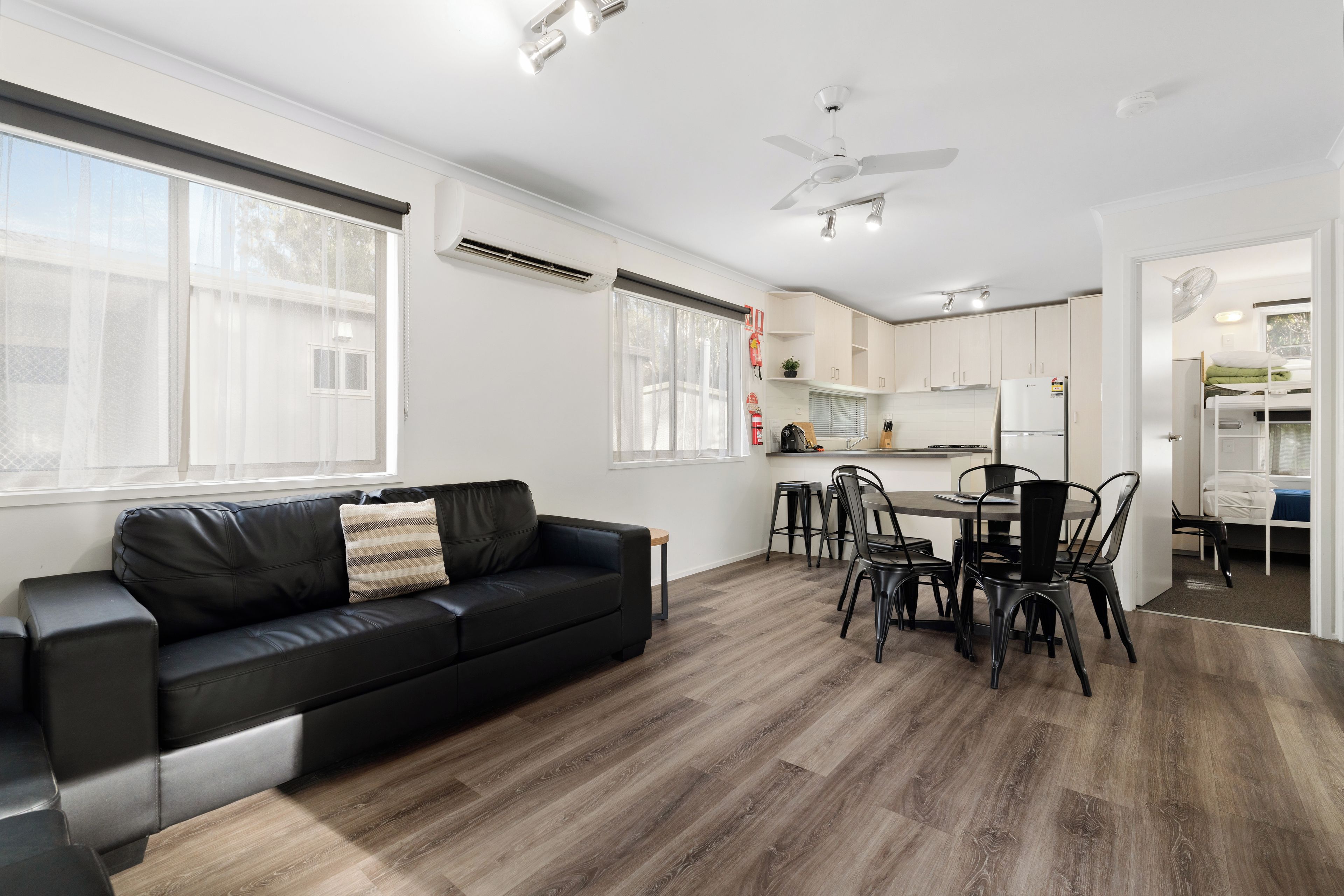 A modern living room with a black leather sofa, a dining area with a table and chairs, and a kitchen with white cabinets and a refrigerator.