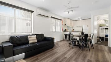 A modern living room with a black leather sofa, a dining area with a table and chairs, and a kitchen with white cabinets and a refrigerator.