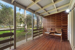 Un espacio de estar al aire libre cubierto con un banco de madera y una mesa, con vista a una zona ajardinada con árboles.