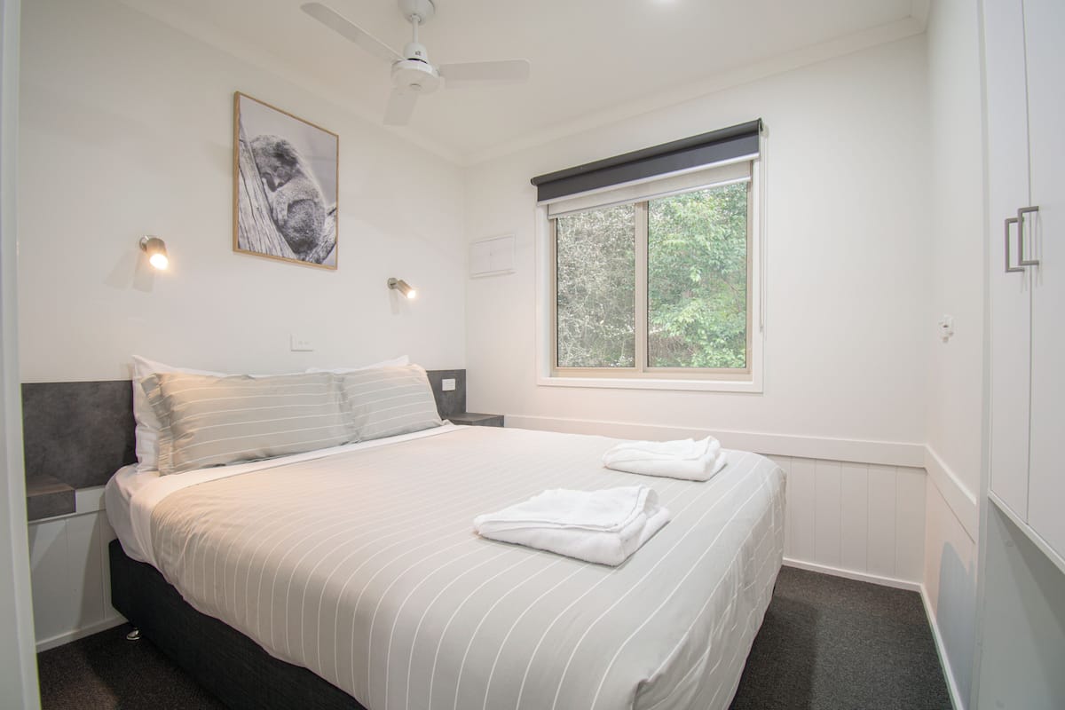 Quarto com cama, janela, ventilador de teto e luminária na parede.
