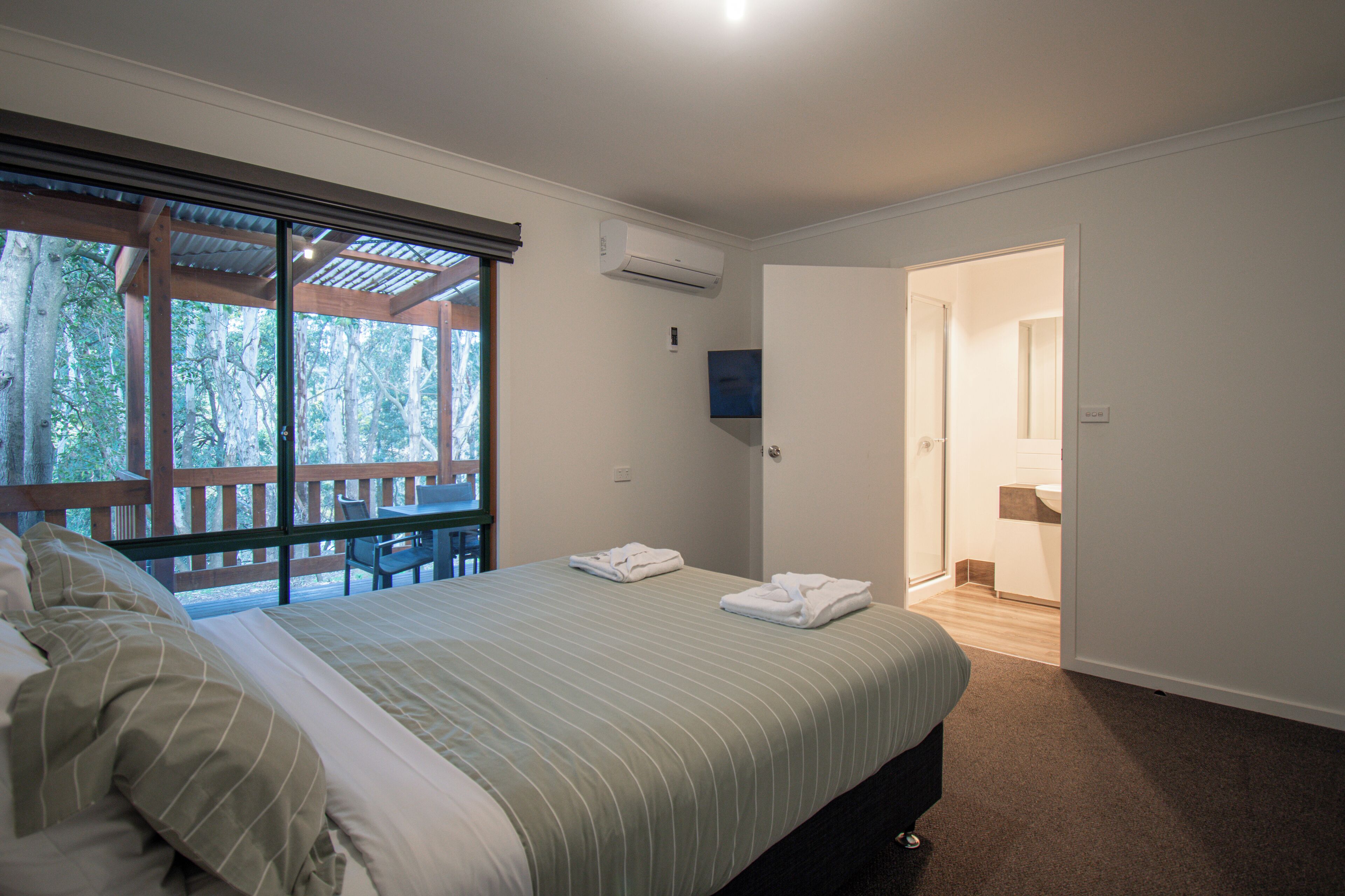 A bedroom with a bed, a window with a view of trees, an air conditioning unit, and a door leading to a bathroom.