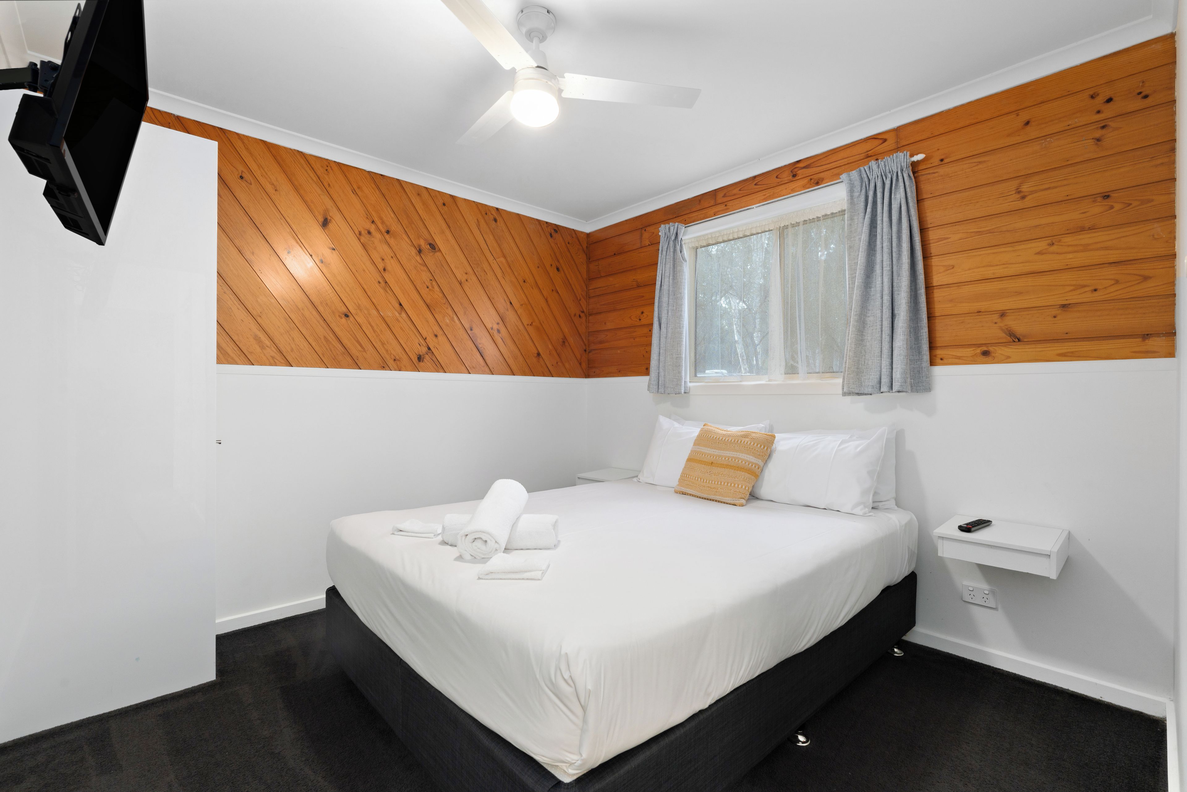Un dormitorio con una cama, una ventana con cortinas, un ventilador de techo y un televisor montado en la pared.