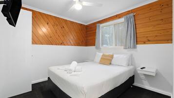 Un dormitorio con una cama, una ventana con cortinas, un ventilador de techo y un televisor montado en la pared.