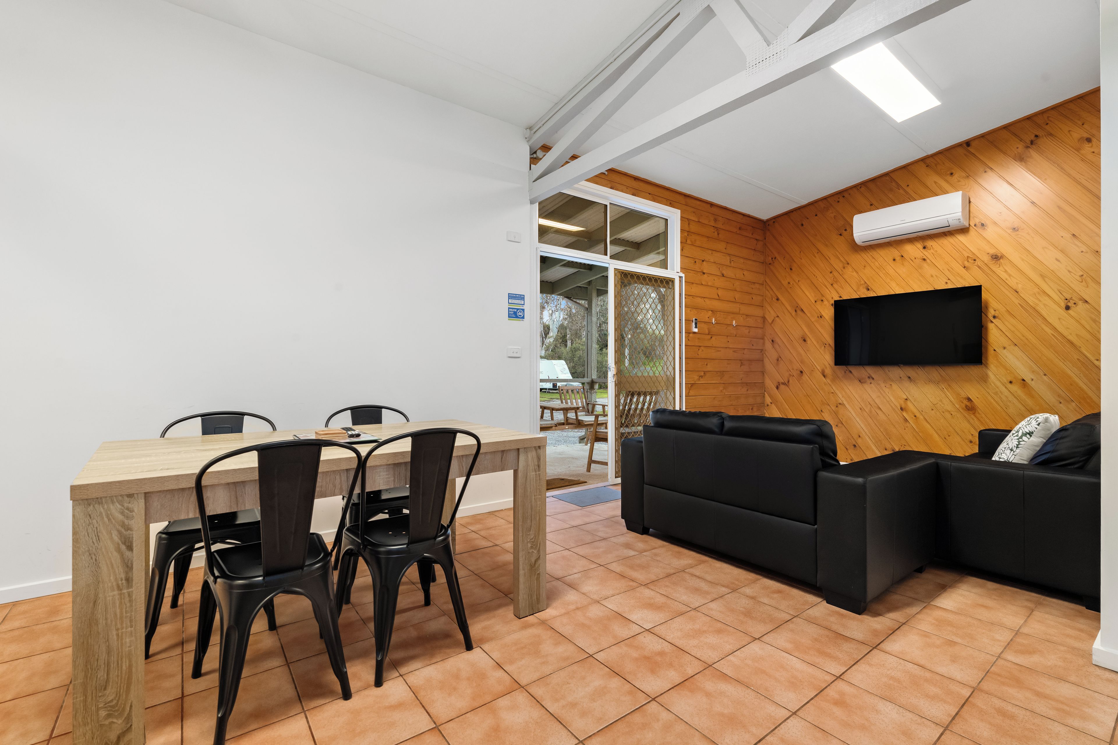 A modern interior with a wooden wall, a flat-screen TV mounted above a black leather sofa, and a dining area with a wooden table and chairs.