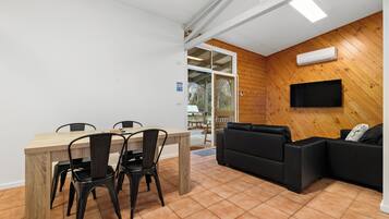 A modern interior with a wooden wall, a flat-screen TV mounted above a black leather sofa, and a dining area with a wooden table and chairs.
