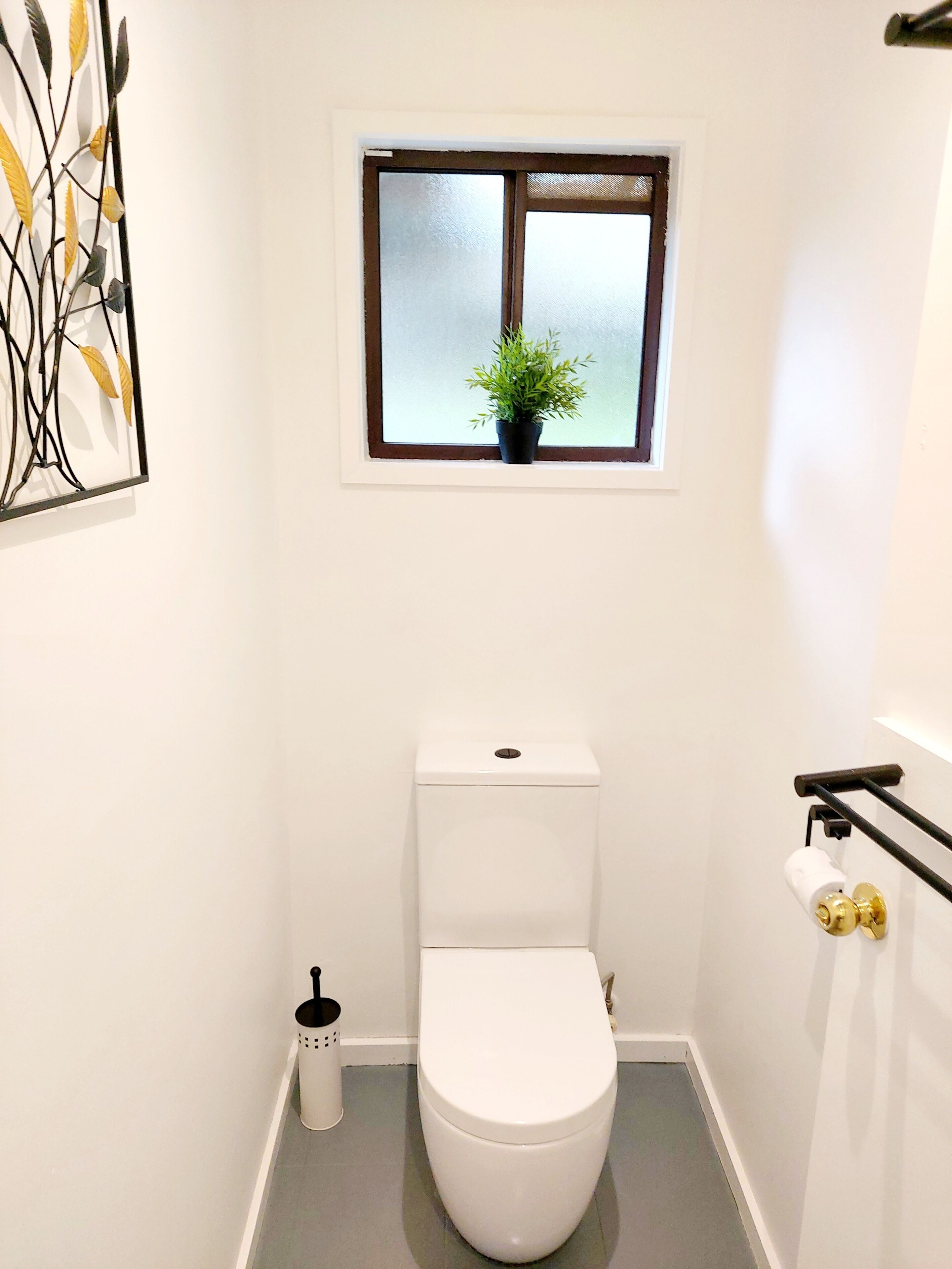 Un baño pequeño y blanco con inodoro, una ventana con planta y un dispensador de papel higiénico en la pared.